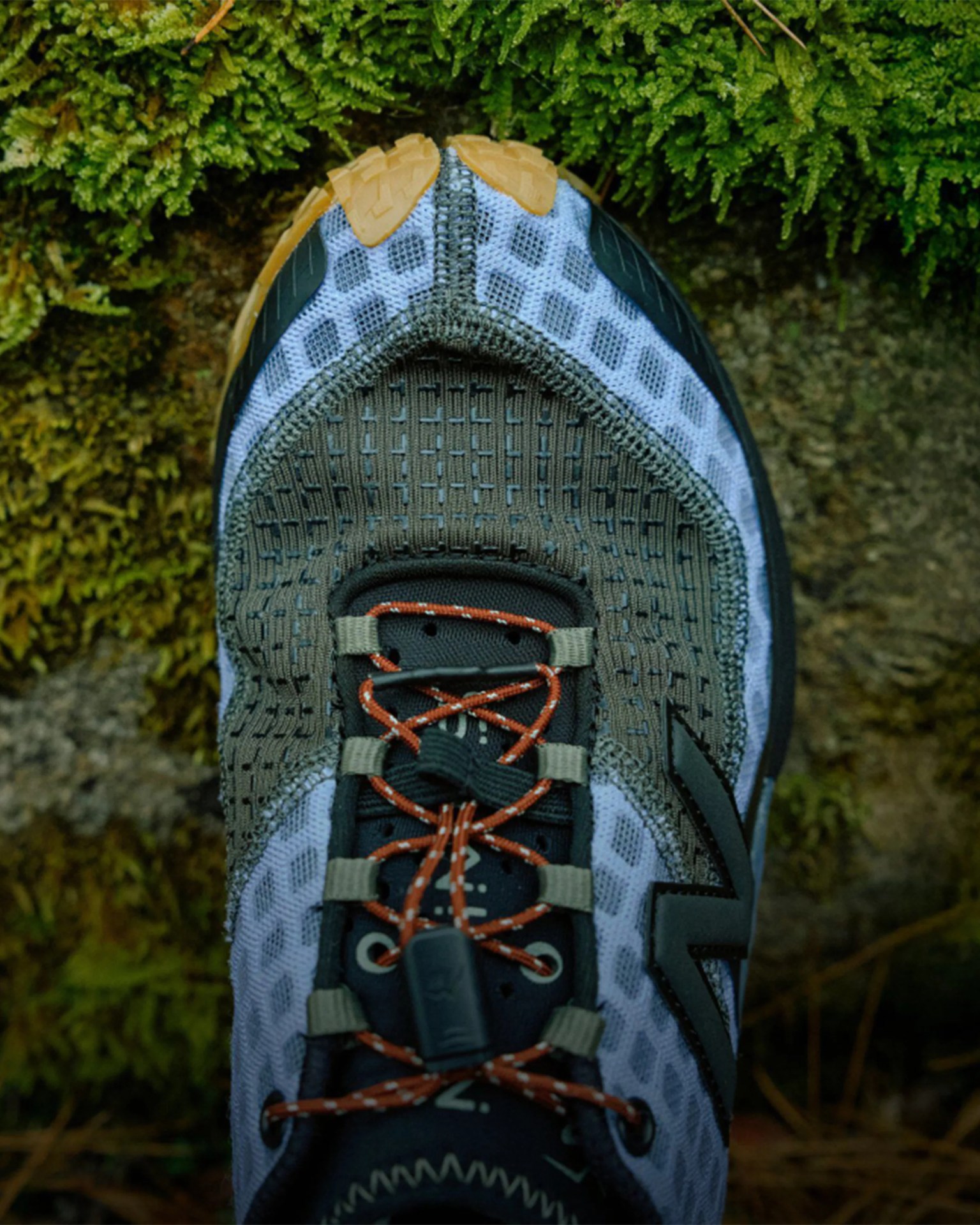Close-up of a gray and black trail running shoe with orange laces on mossy ground.