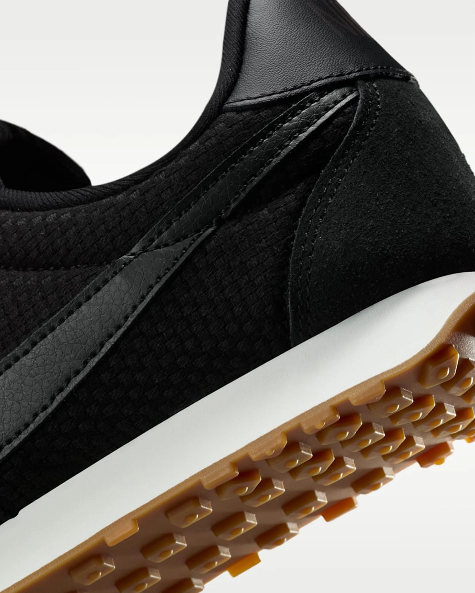 Close-up of black sneaker with textured fabric, leather accents, white midsole, and gum rubber outsole with hexagonal tread.