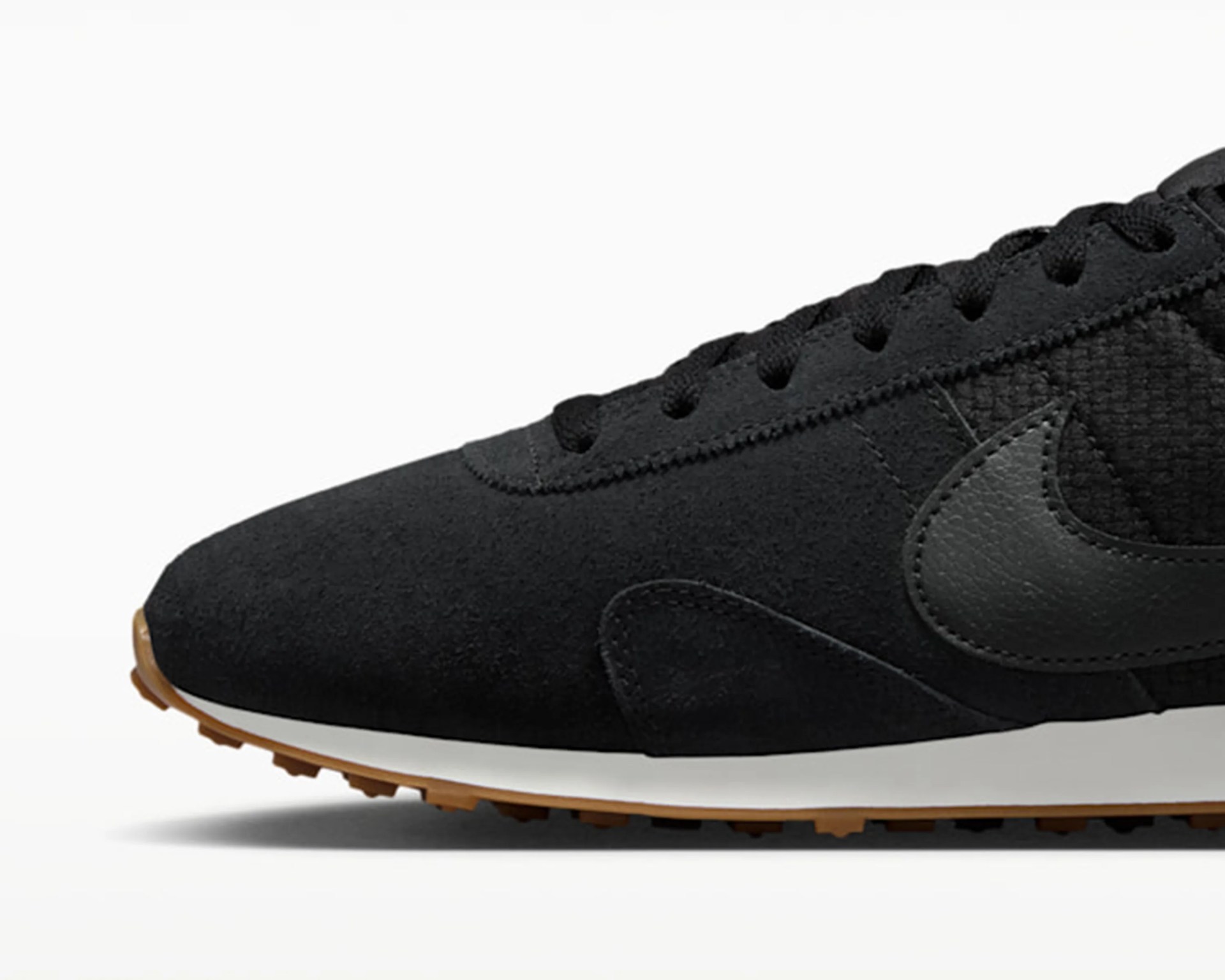 Close-up of black Nike sneaker with suede and mesh upper, black leather swoosh, white midsole, and gum rubber outsole.