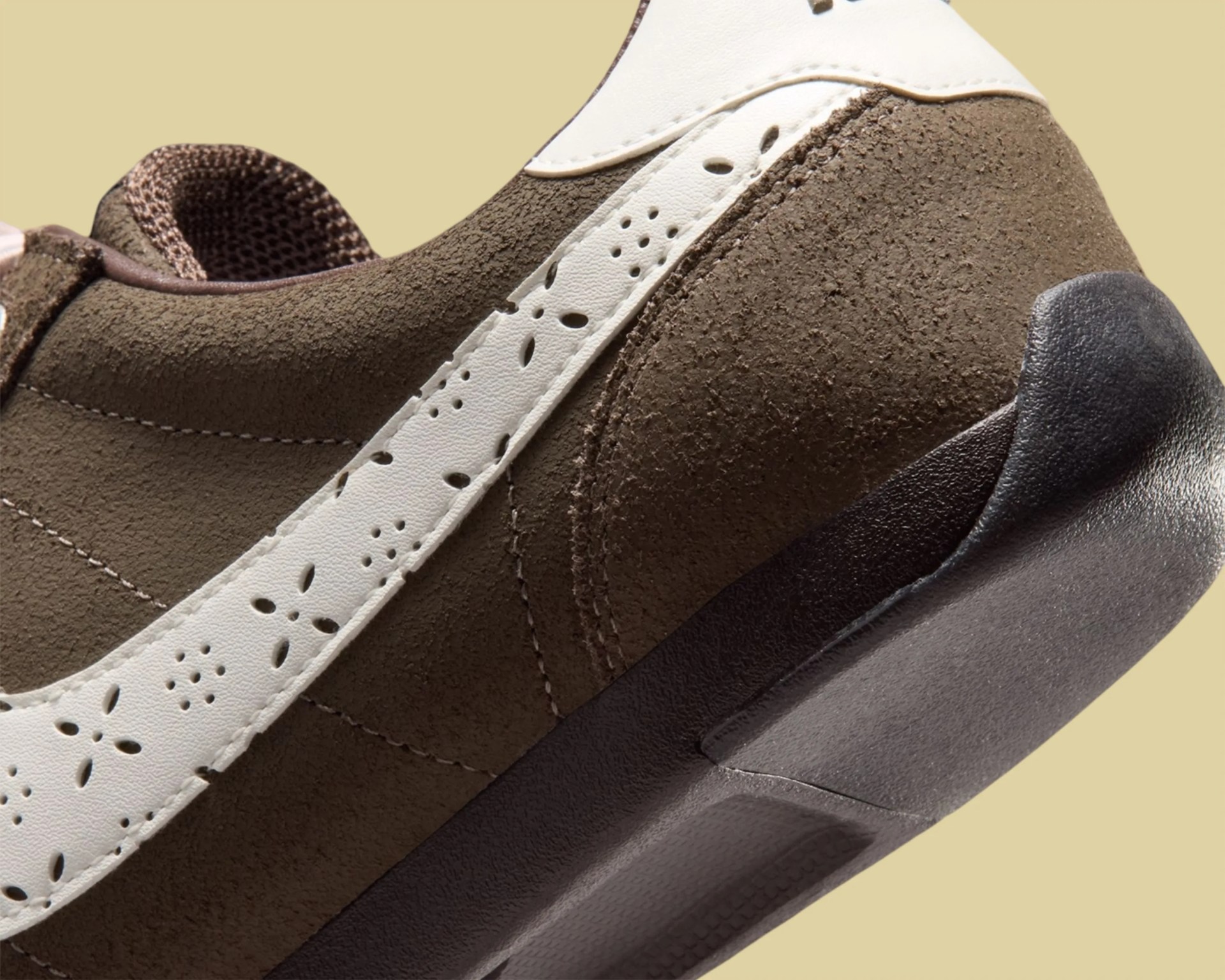 Close-up of brown suede sneaker with white perforated leather stripe and black rubber sole.