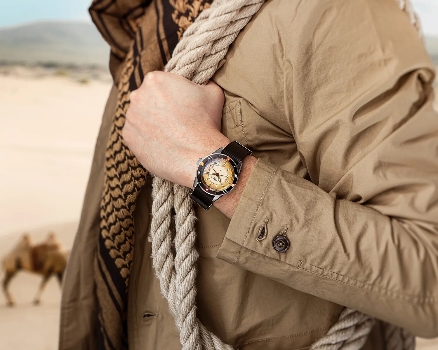 Wristwatch with a beige dial and black strap worn on a wrist holding a thick rope, with a camel in the desert background.