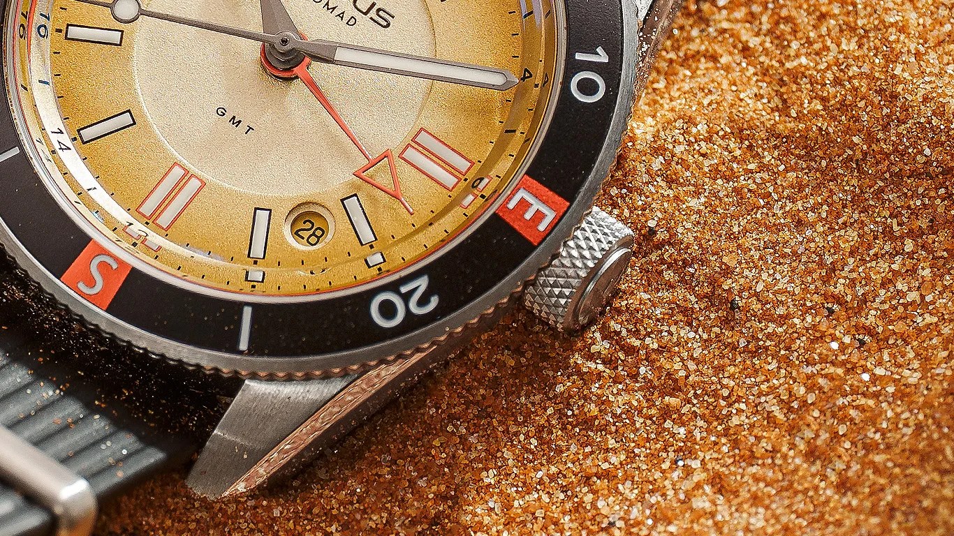 Close-up of a gold-toned wristwatch with black bezel and red accents partially buried in golden sand.