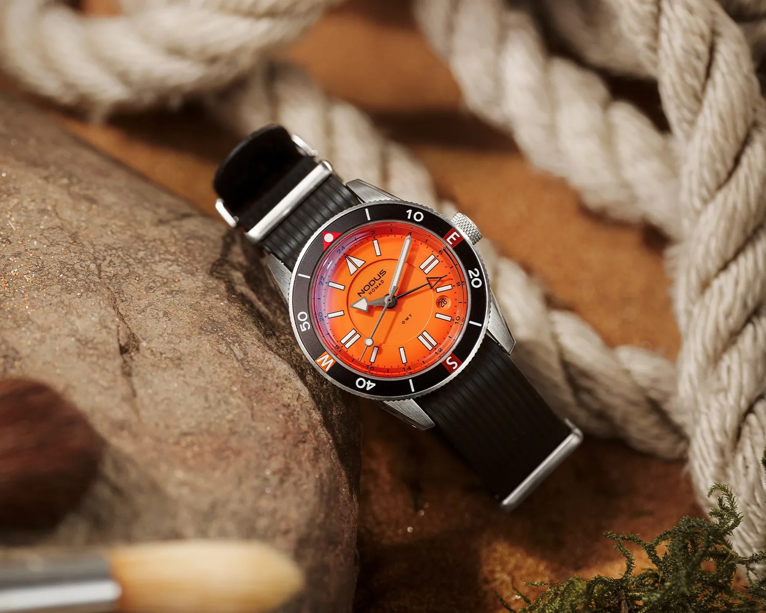 Stainless steel watch with bright orange dial, black bezel marked with compass directions, and black textured strap on a rock surface.