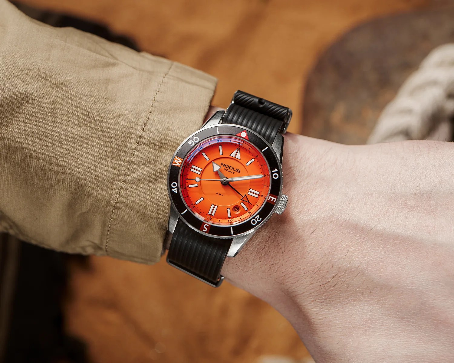Wristwatch with bright orange dial, black bezel marked with compass directions, and black ribbed strap worn on a wrist.