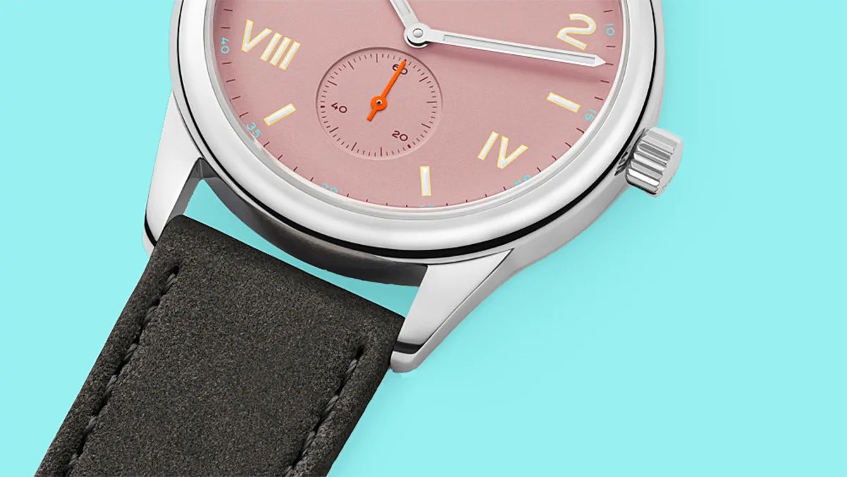 Close-up of a wristwatch with a pink dial, gold Roman numerals, silver case, and black leather strap on a turquoise background.