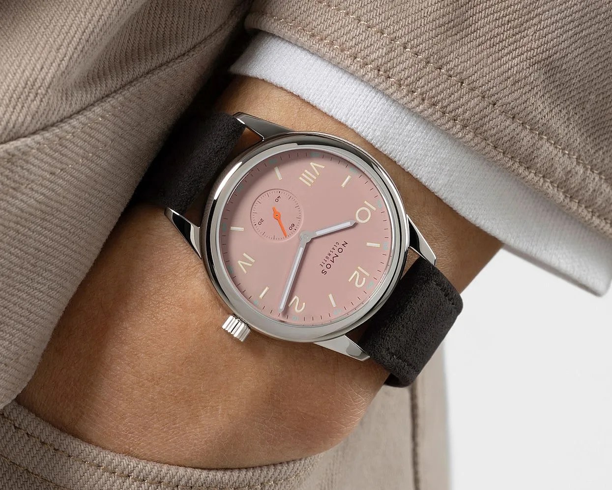 Wristwatch with a pink dial, silver case, black suede strap, and beige numerals worn on a wrist with beige clothing.