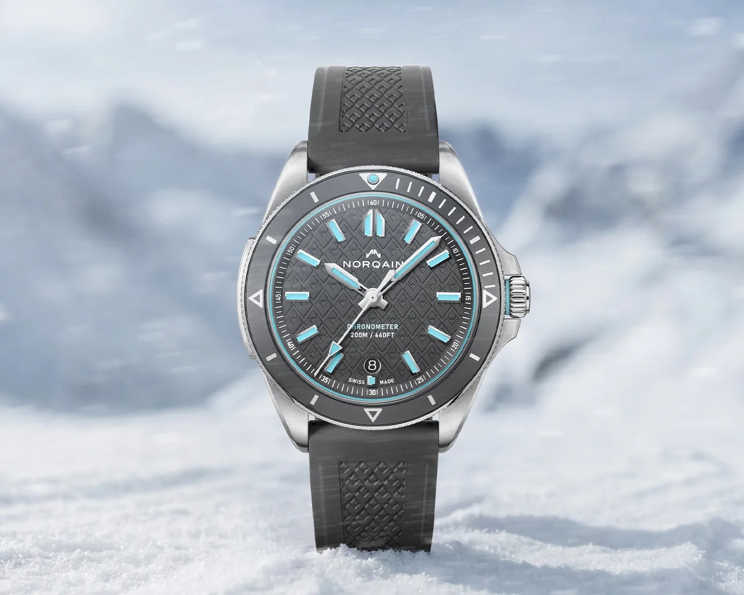 Silver Norqain chronometer watch with gray textured dial, light blue markers, and black rubber strap on snow.