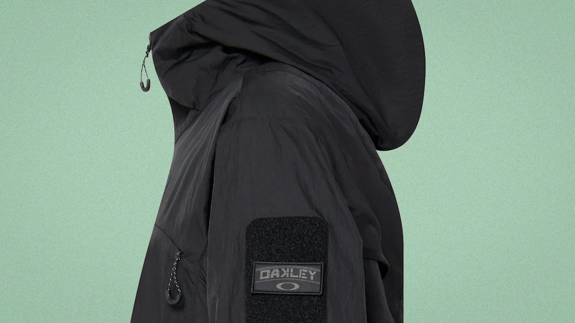 Black Oakley jacket with hood and zippered pocket on the sleeve against a green background.