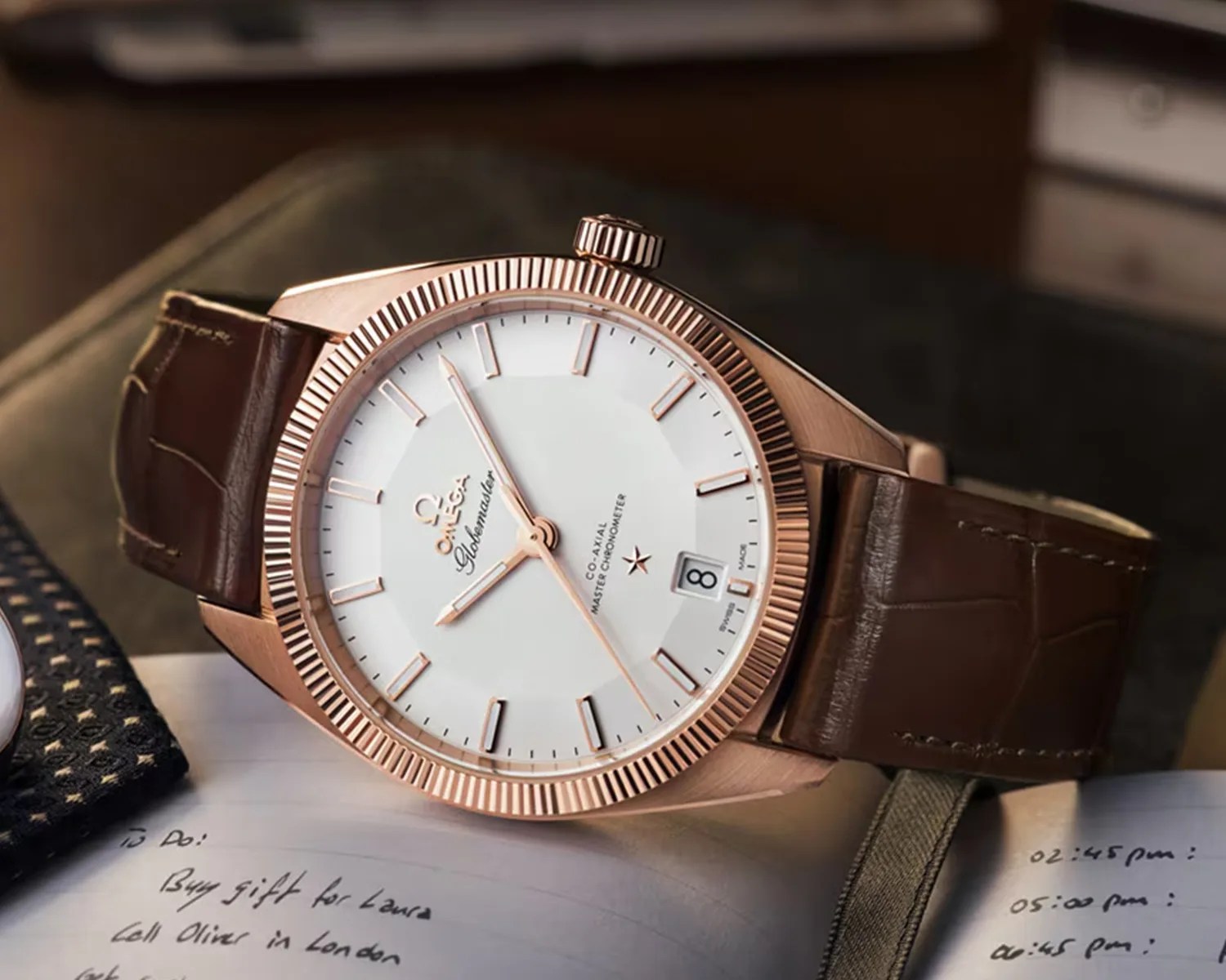 Rose gold Omega Globemaster wristwatch with white dial and brown leather strap resting on an open notebook.