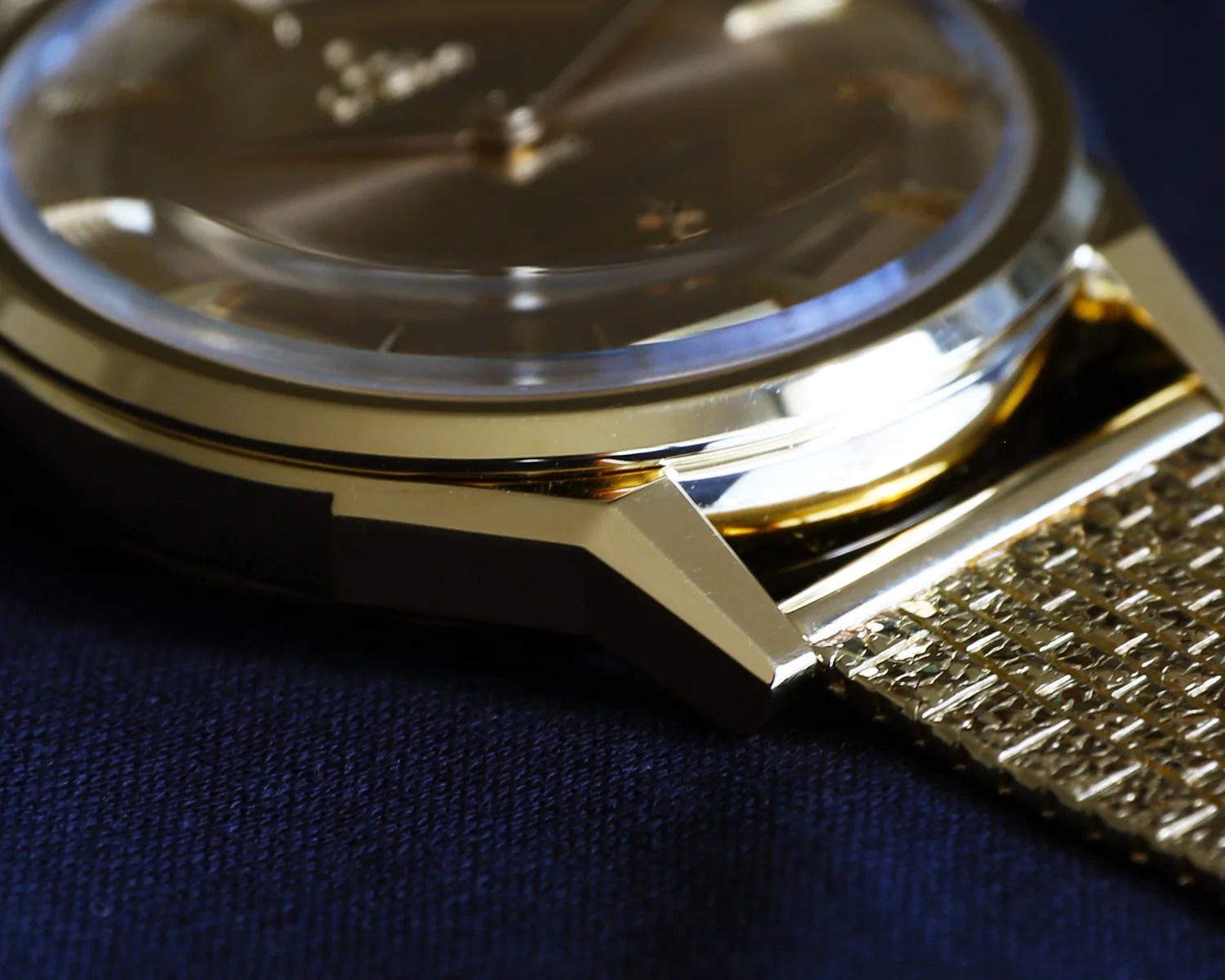 Close-up of a gold watch case with a textured gold mesh strap on a dark fabric background.