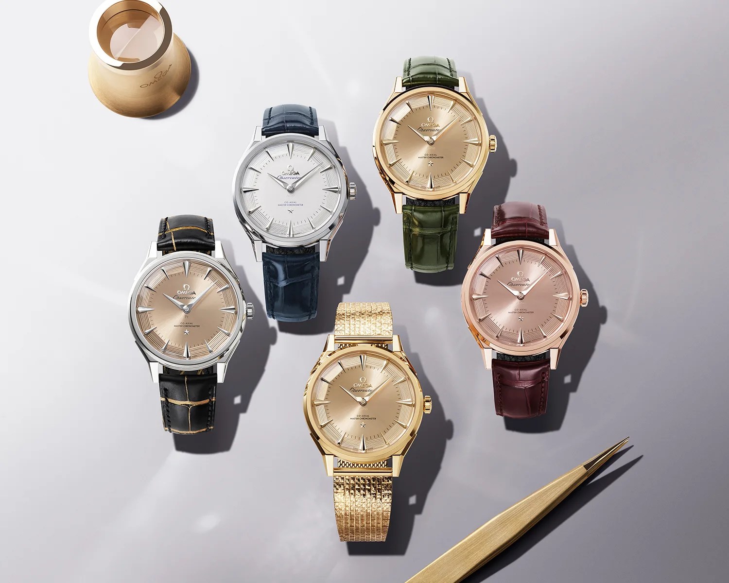 Five Omega wristwatches with leather and metal bands in black, blue, green, burgundy, and gold on a gray surface with gold watchmaking tools.