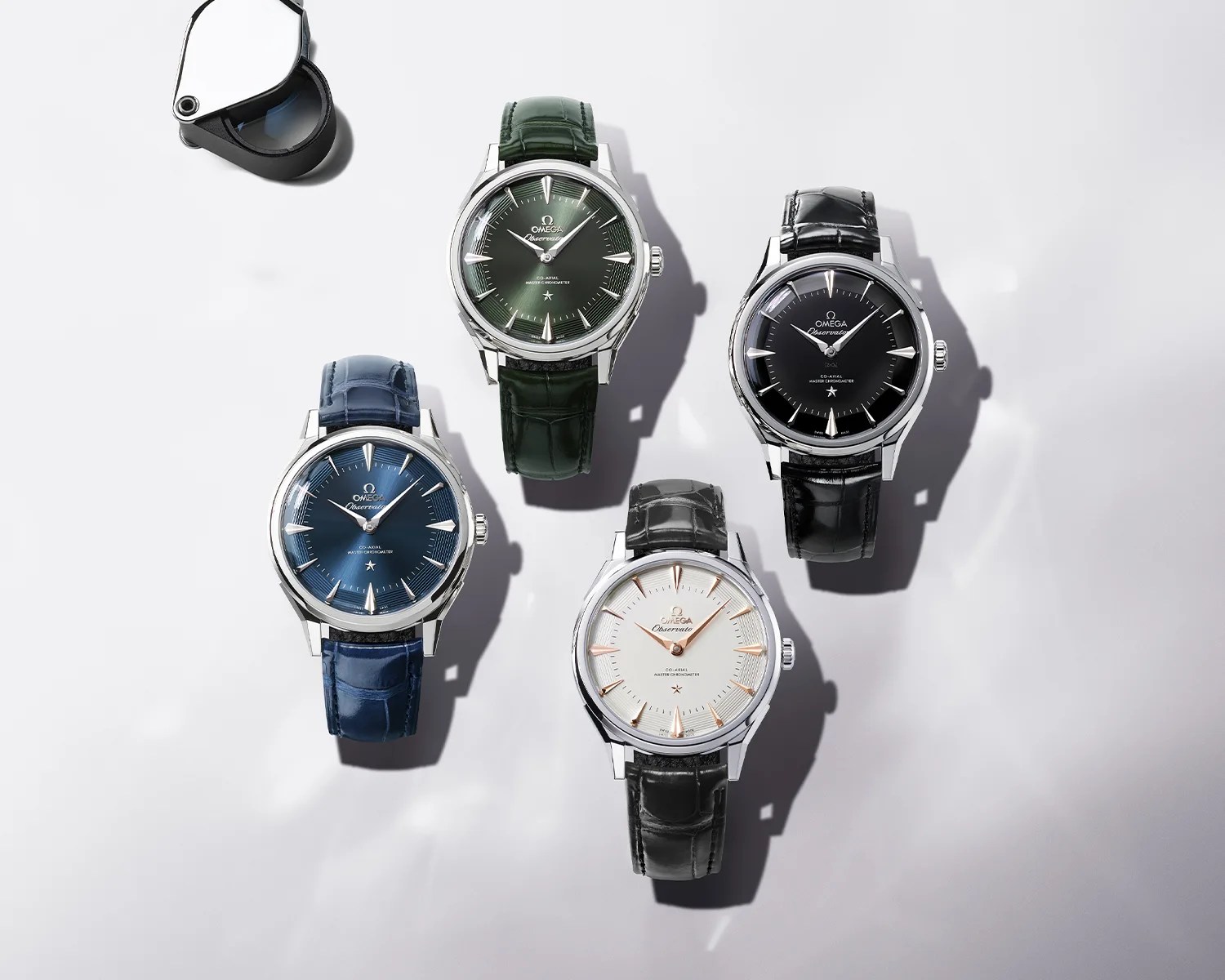 Four Omega wristwatches with leather straps in green, black, blue, and black with white dial, arranged on a white surface with a watch loupe nearby.
