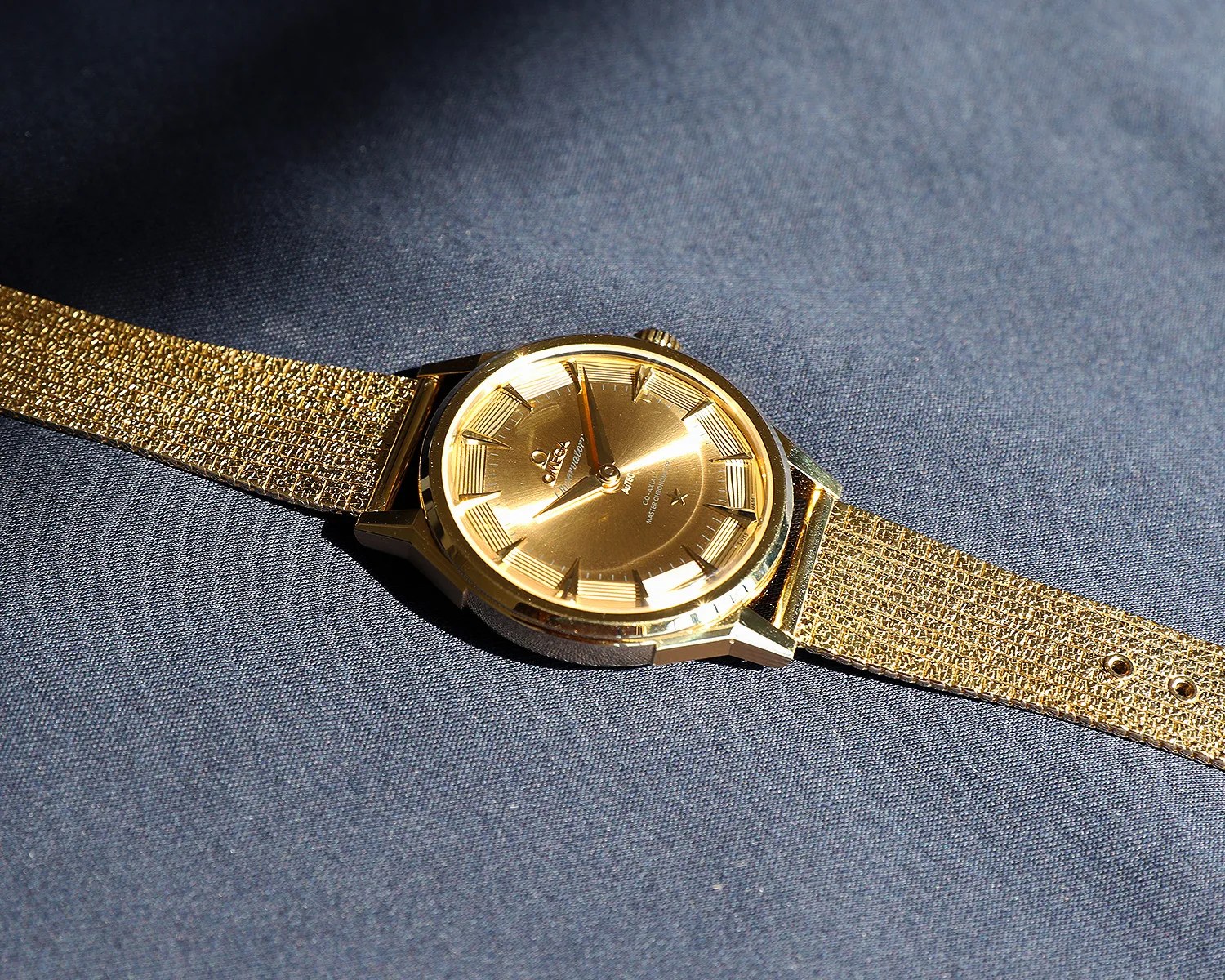 Gold Omega wristwatch with textured gold mesh strap on dark fabric.