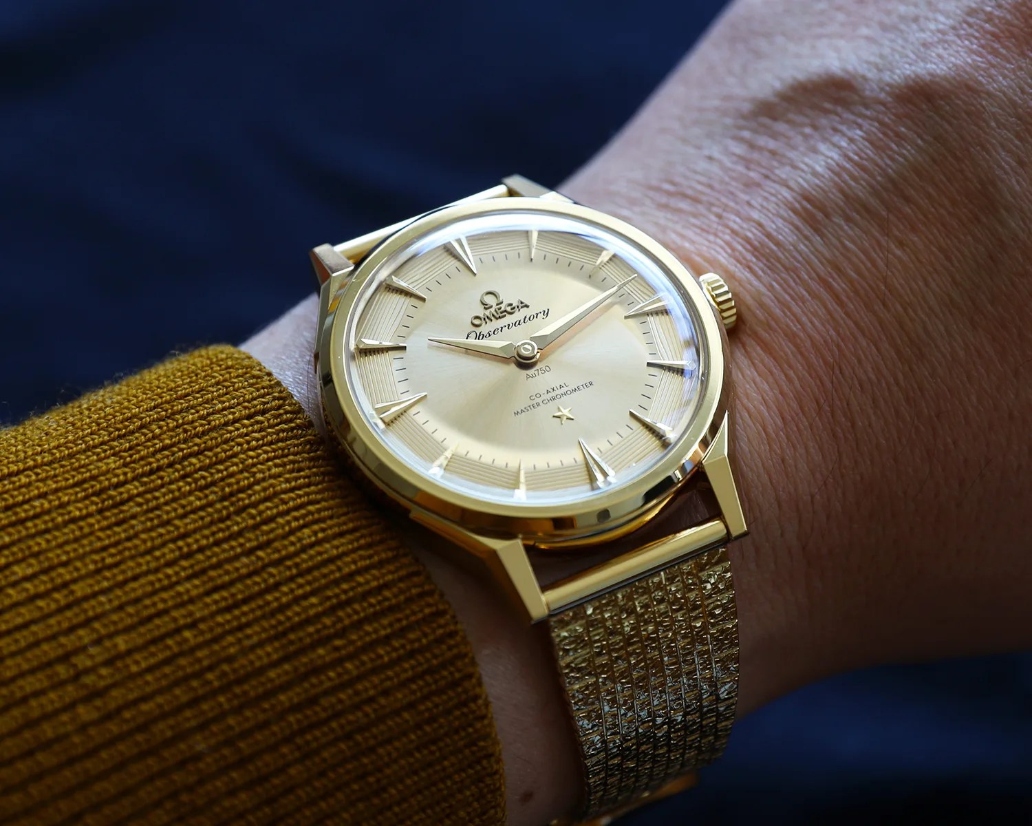 Gold Omega Observatory wristwatch with textured gold mesh strap worn on a wrist with a mustard yellow sweater.