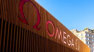 Large red Omega logo and text mounted on a vertical wooden slat wall with a building and blue sky in the background.