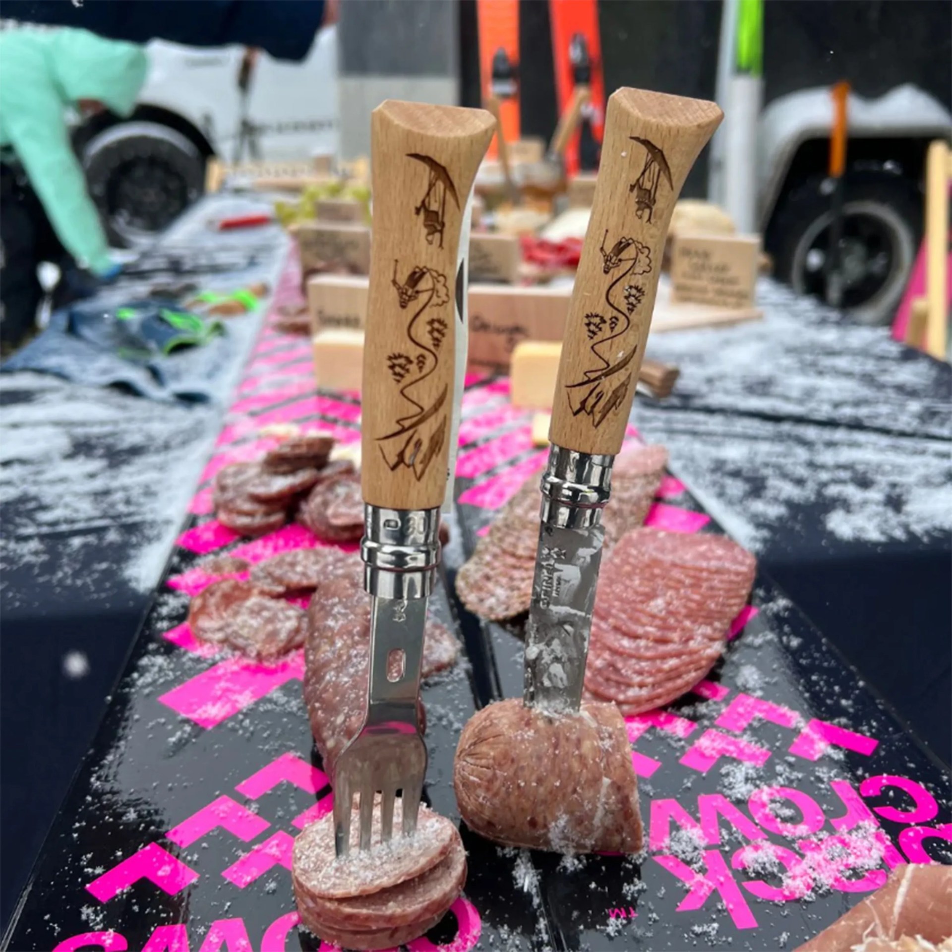 Two wooden-handled knives with engraved dragon designs stuck into slices of cured meat on a black surface with pink text.
