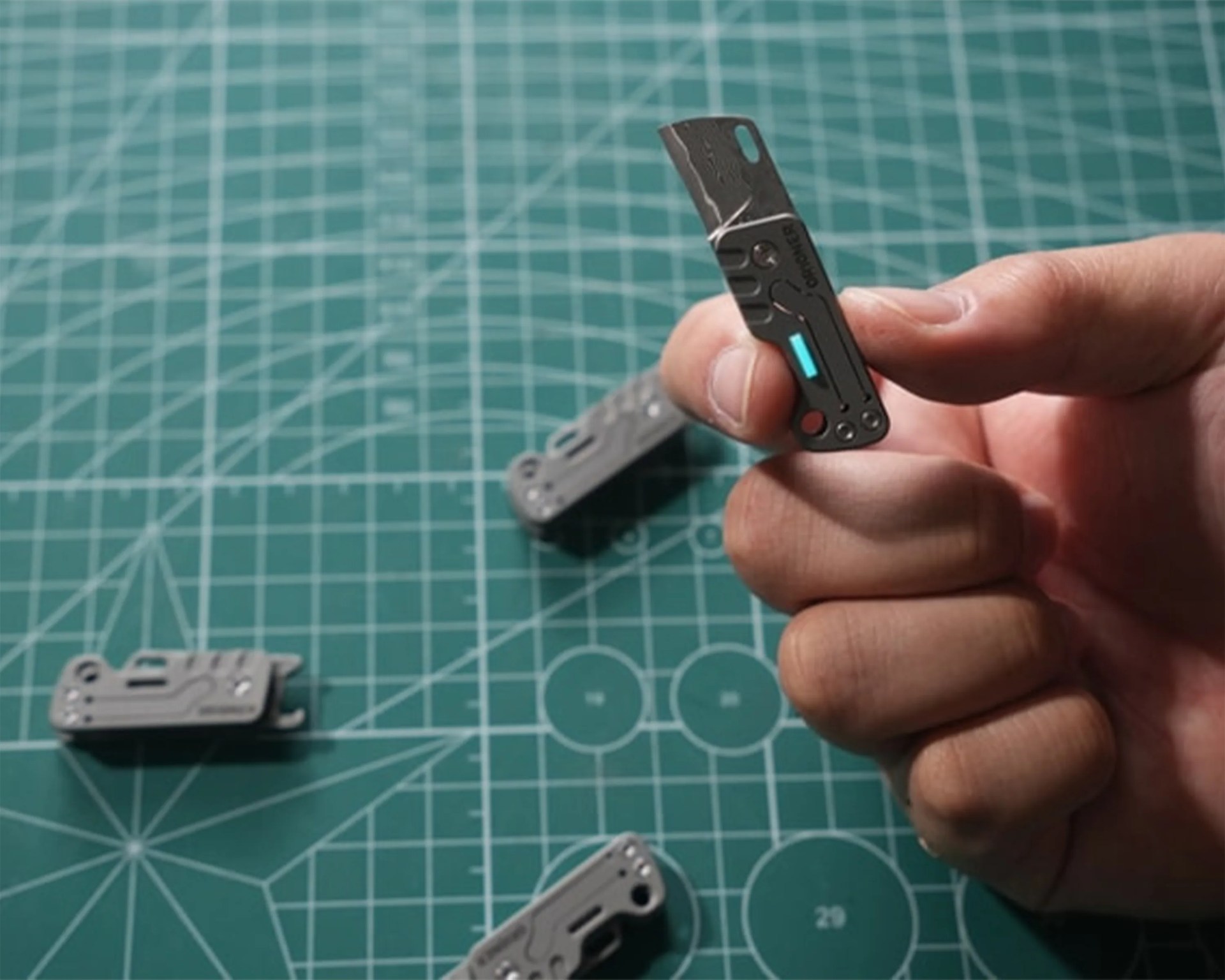 Hand holding a small black utility knife with a blue accent above a green cutting mat with grid lines and other similar knives.