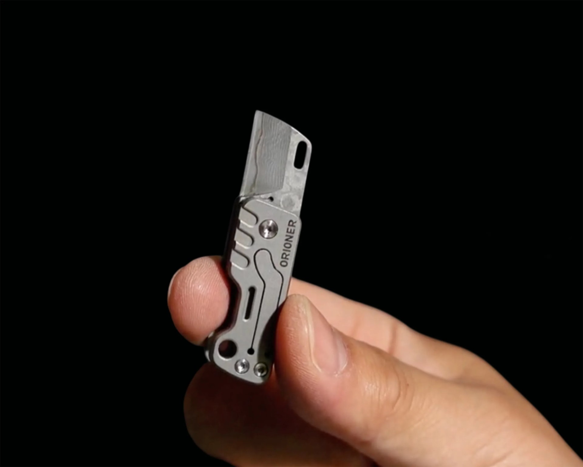 Hand holding a small silver folding knife with "ORIONER" engraved on the handle against a black background.