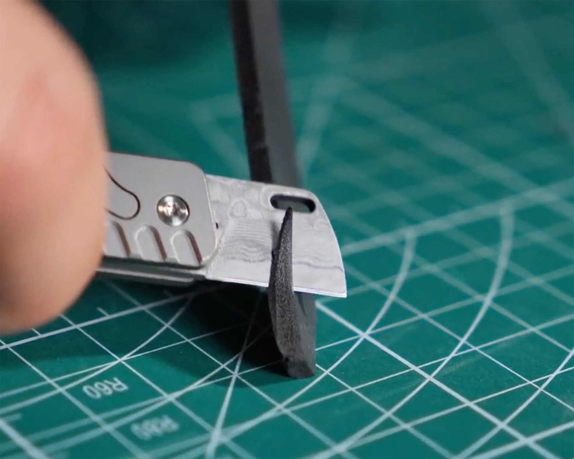 Utility knife blade cutting a thin black strip on a green cutting mat with white grid lines.