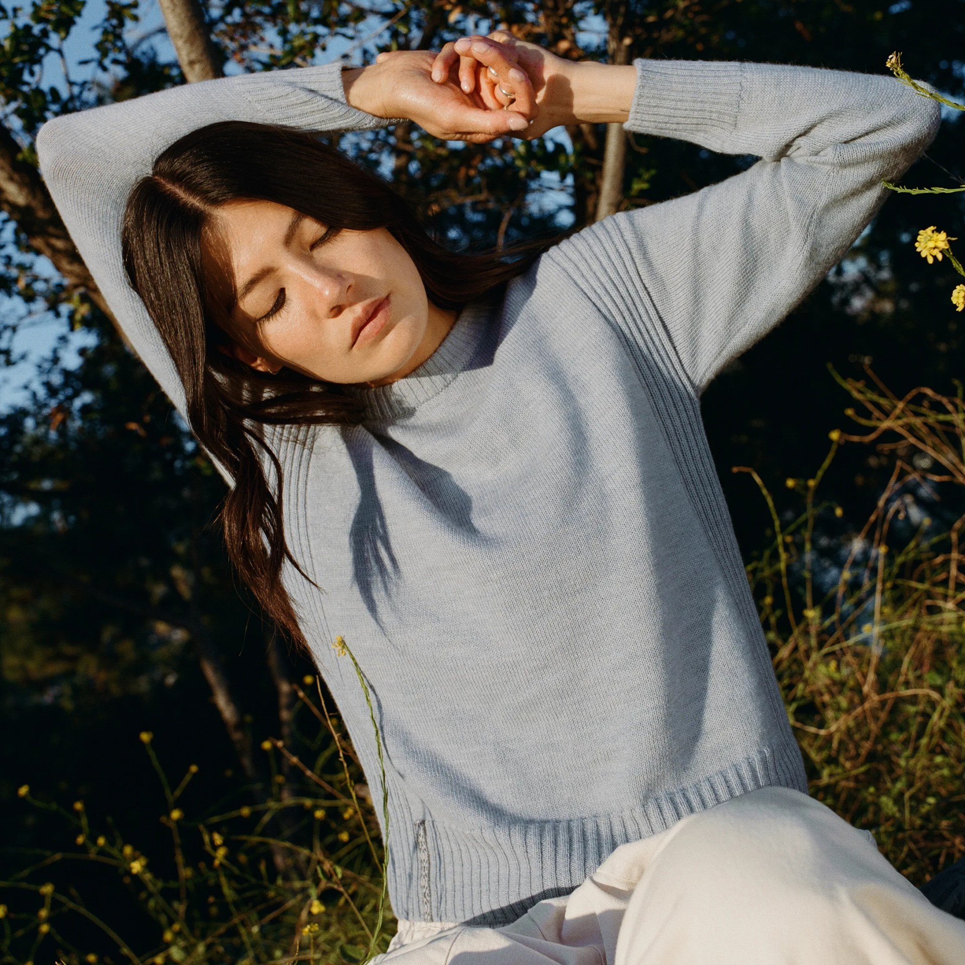Woman wearing a light gray ribbed knit sweater with long sleeves outdoors among greenery.