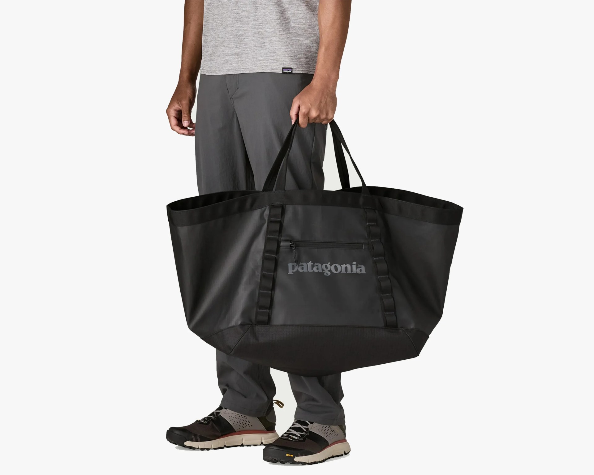 Large black Patagonia tote bag with front zipper pocket held by a person wearing gray pants and black sneakers.