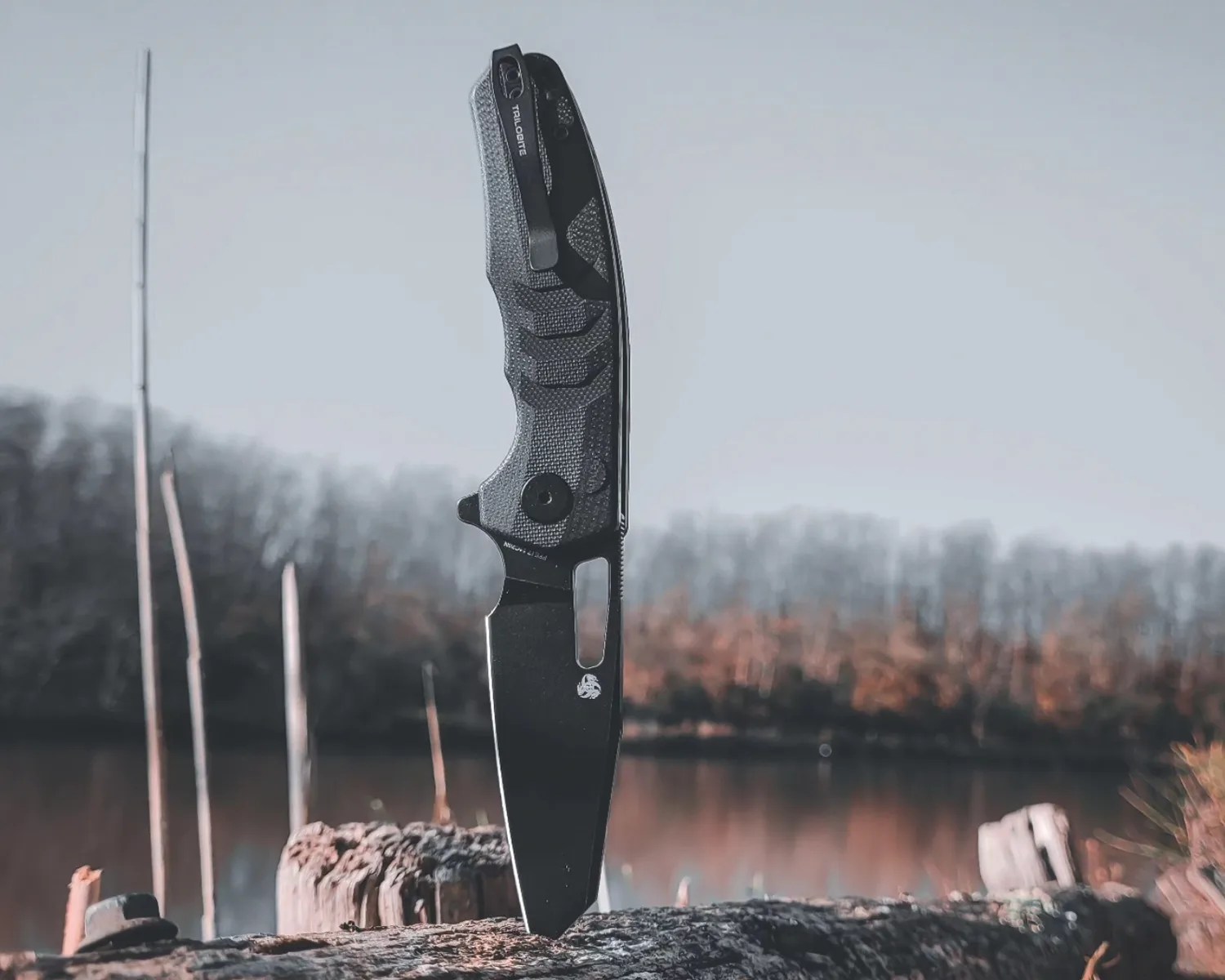 Black folding knife with textured handle standing blade-down on a log near a lake.