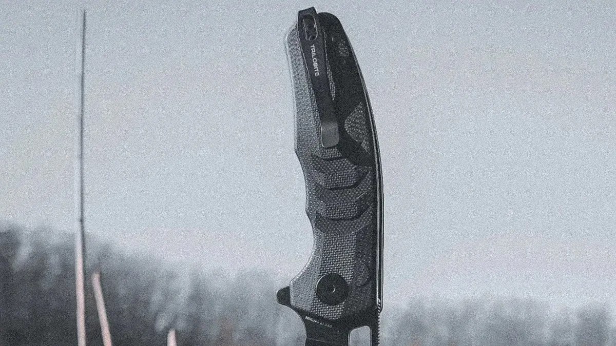 Black textured folding knife handle with a clip, held upright against a blurred outdoor background.