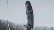Black textured folding knife handle with a clip, held upright against a blurred outdoor background.