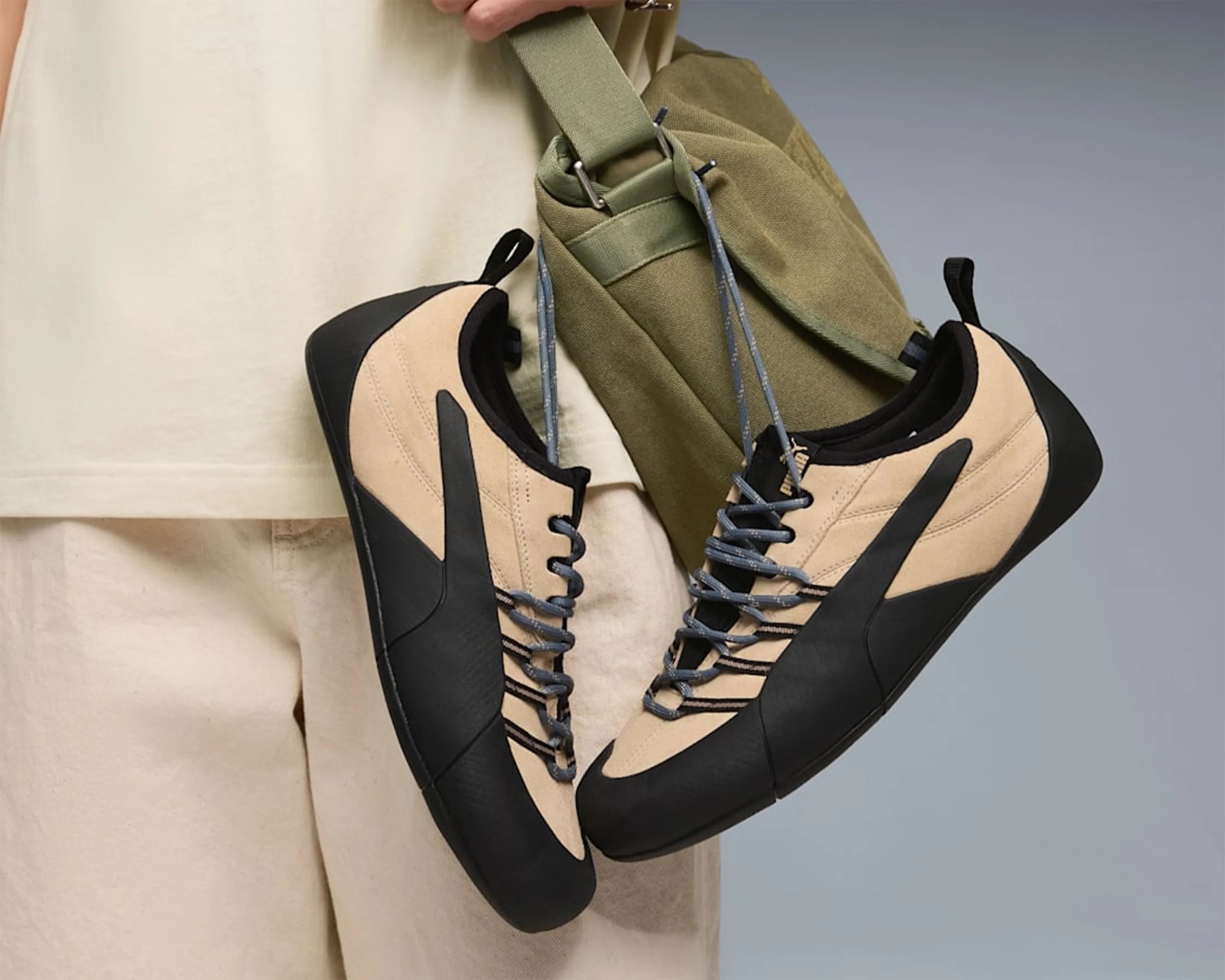 Pair of beige and black Puma sneakers with gray laces hanging from an olive green bag strap.
