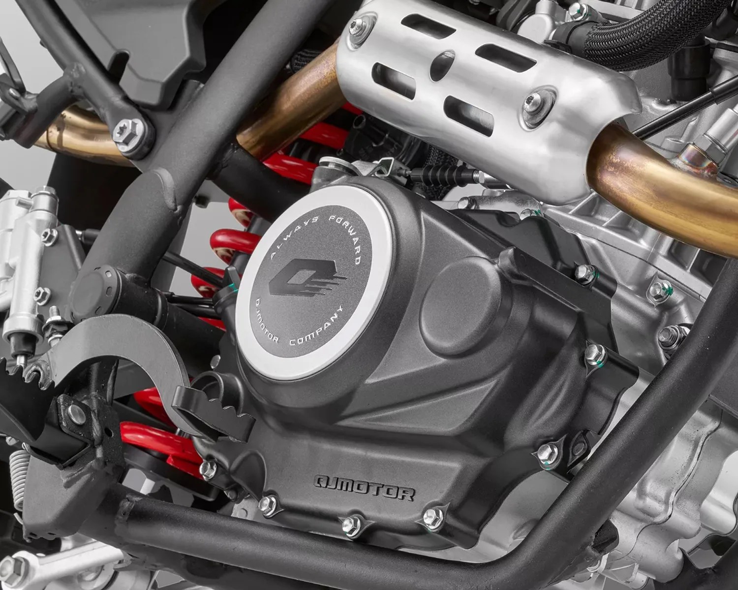 Close-up of a black QJMotor engine casing with silver circular cover and red coil spring in a motorcycle frame.