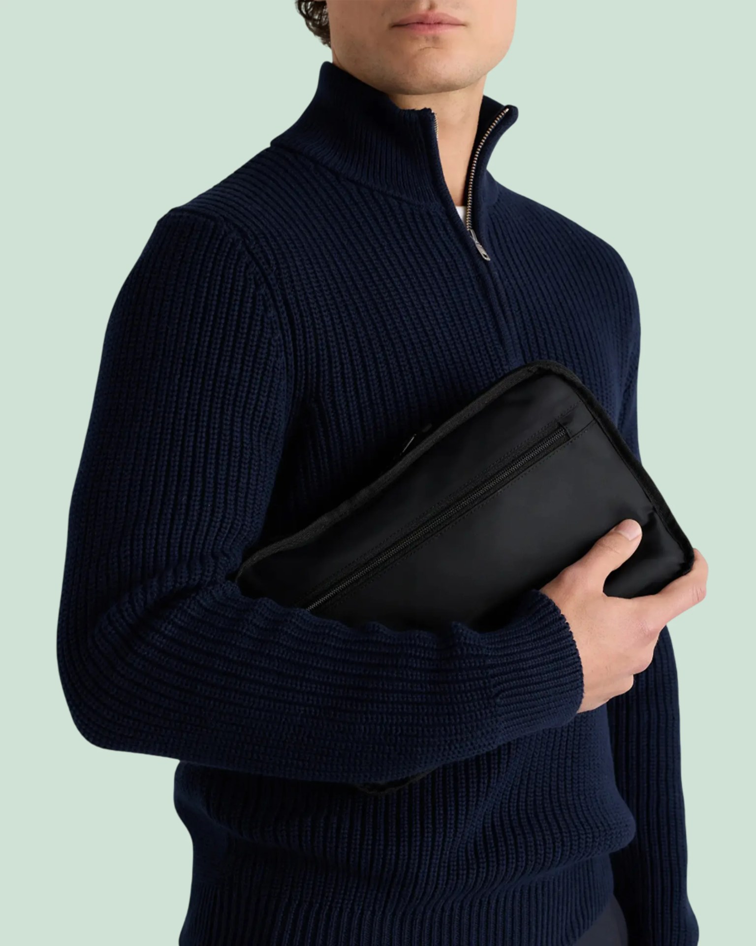 Person wearing a navy ribbed half-zip sweater holding a black zippered pouch in a hand.