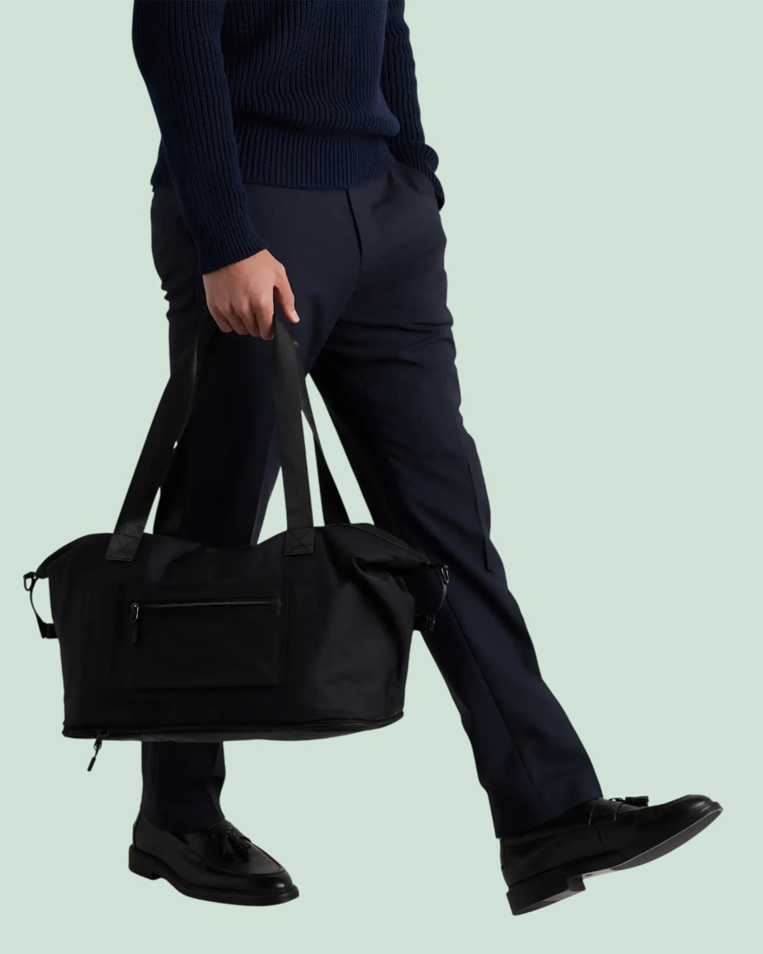 Person wearing navy pants and black loafers holding a black duffel bag with zipper pockets.
