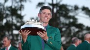 Man in a green jacket holding a silver and wooden trophy with a building model on top.