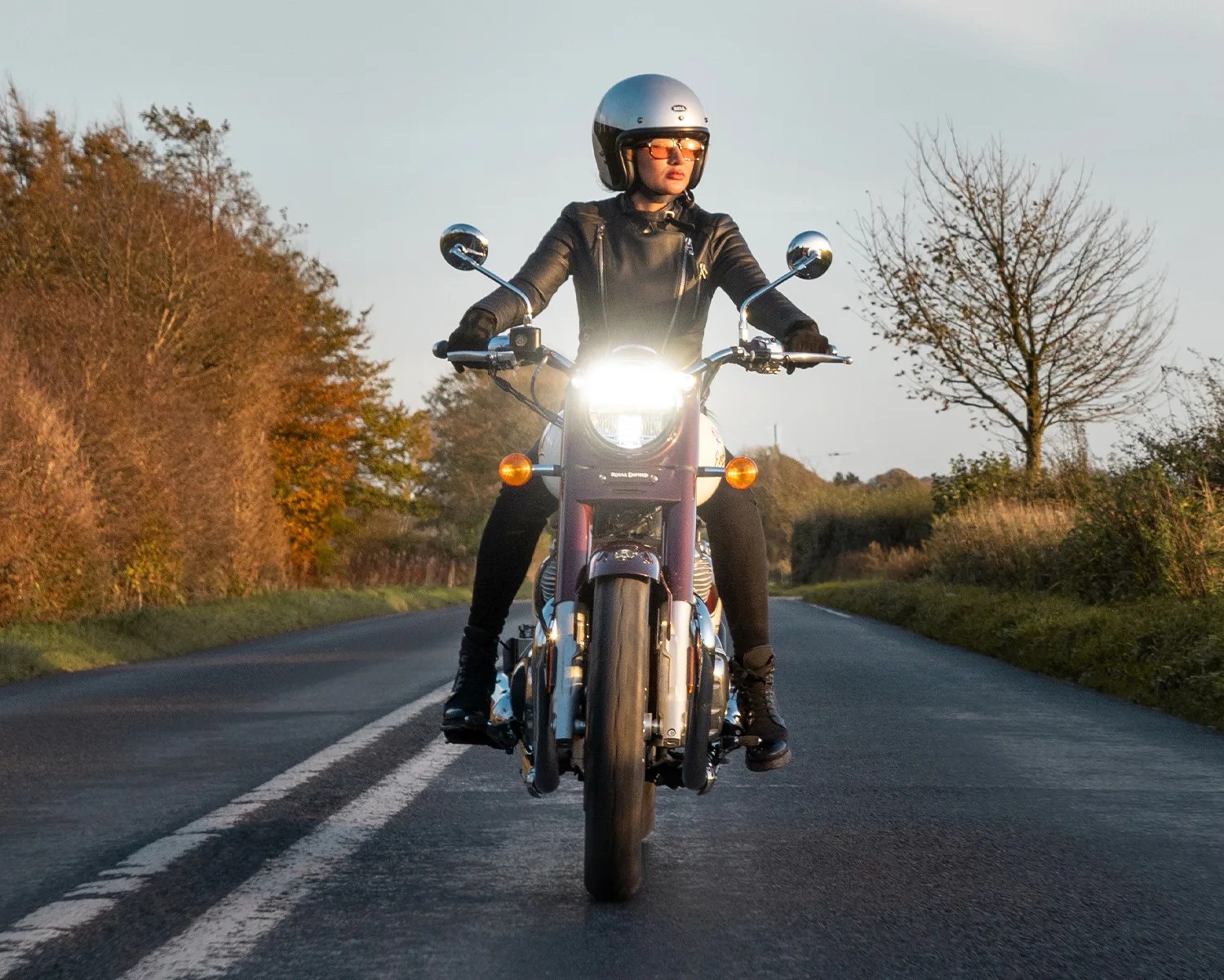 Person wearing a silver helmet and black leather jacket riding a motorcycle with headlight on on a rural road.