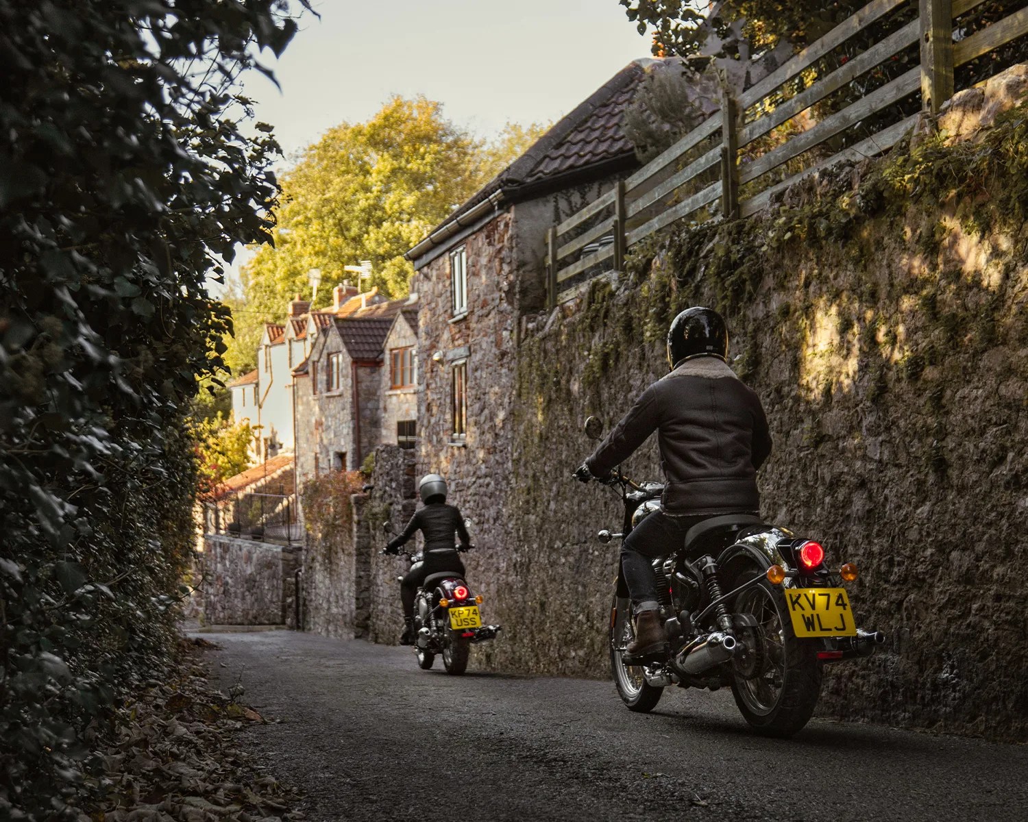 Two motorcyclists wearing helmets riding on a narrow road beside stone walls and houses.