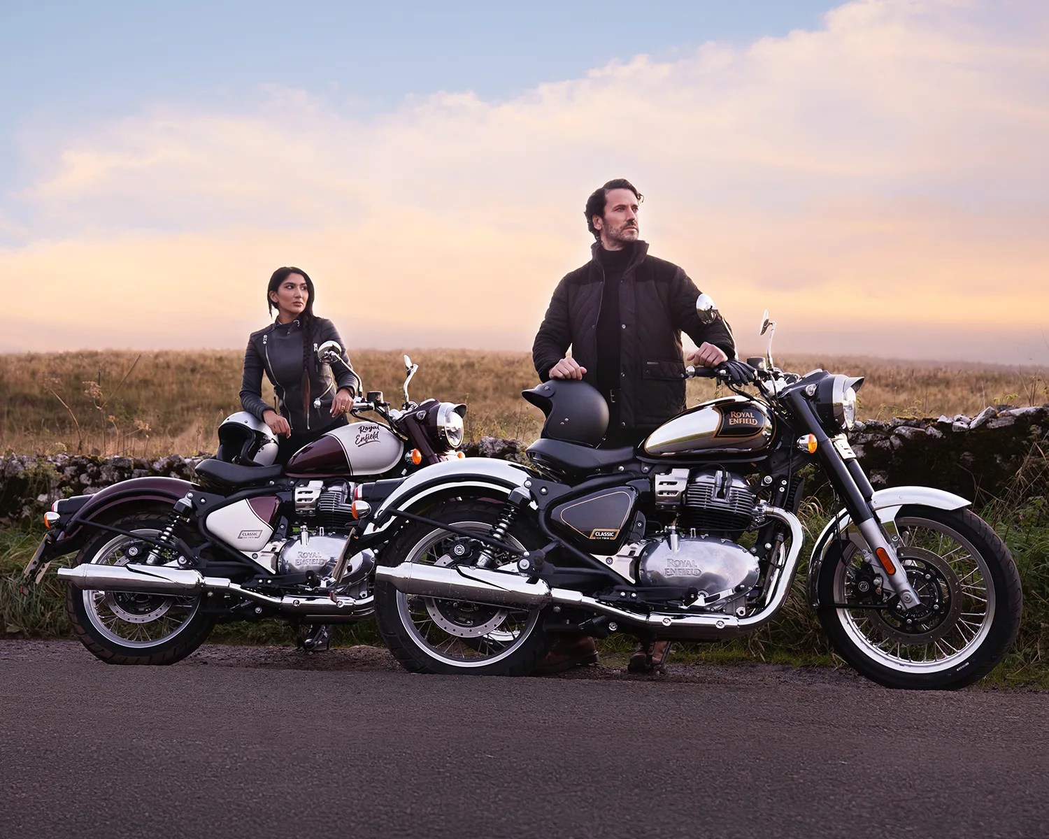 Two Royal Enfield Classic motorcycles parked on a road with riders in black jackets standing beside them at sunset.