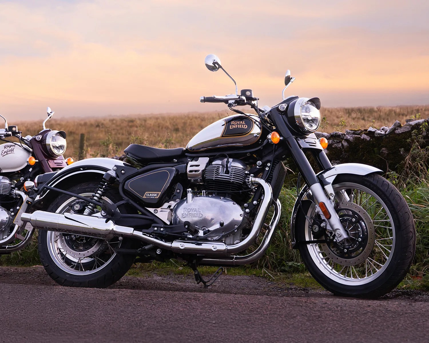 Black and silver Royal Enfield Classic 650 Twin motorcycle parked on a road with a grassy field and stone wall in the background.