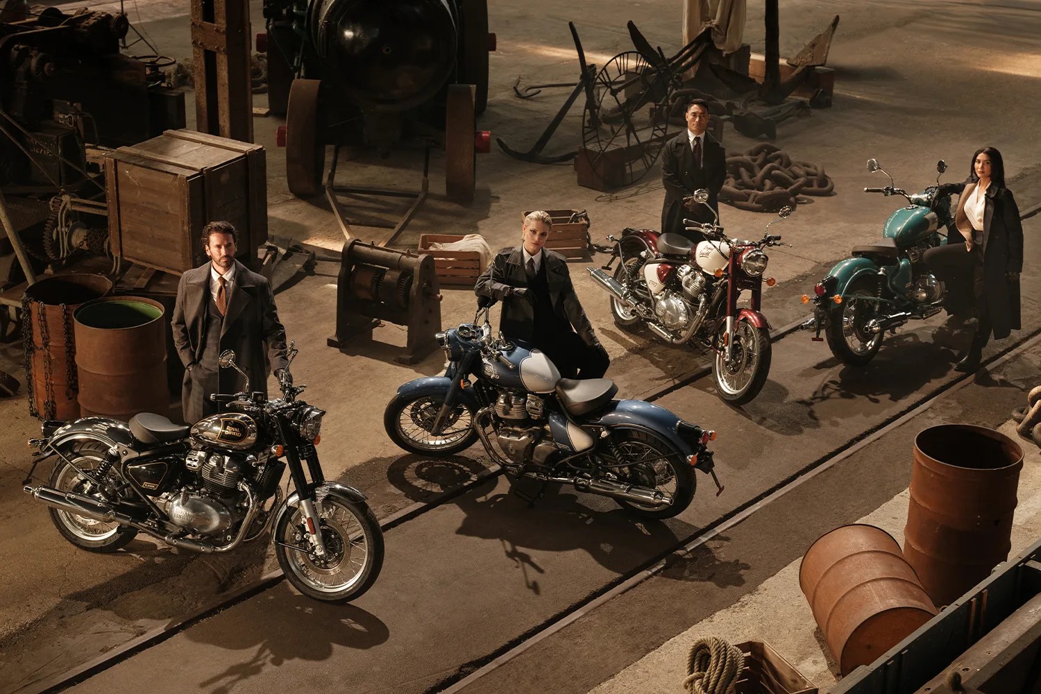 Four people standing or sitting near four Royal Enfield motorcycles in a rustic industrial setting with barrels and wooden crates.