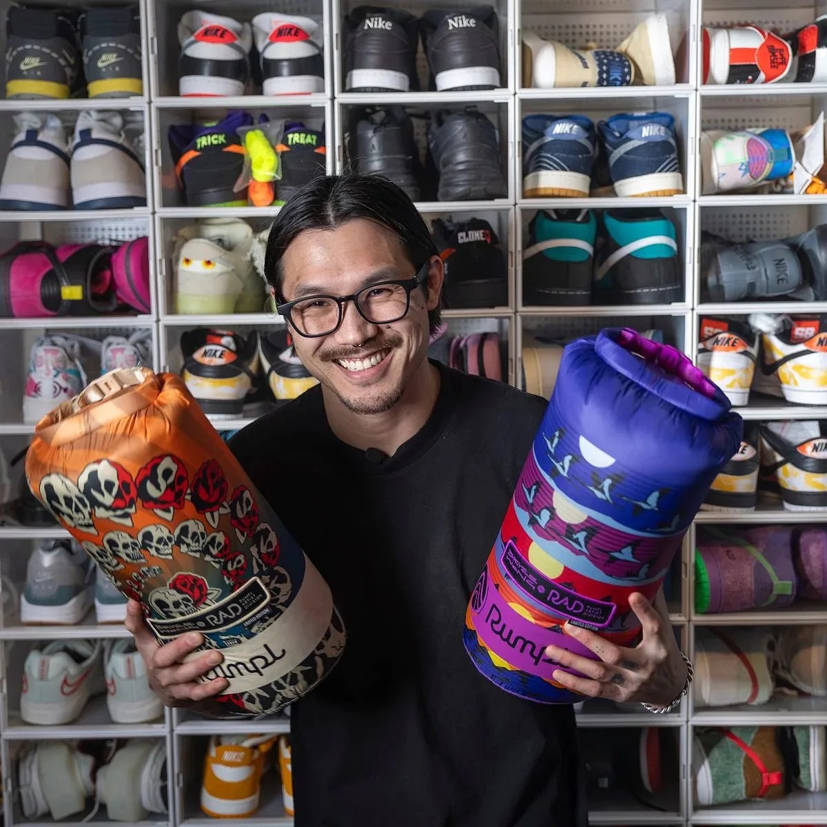 Person wearing glasses holding two colorful rolled-up sleeping bags in front of a wall of cubby shelves filled with sneakers.
