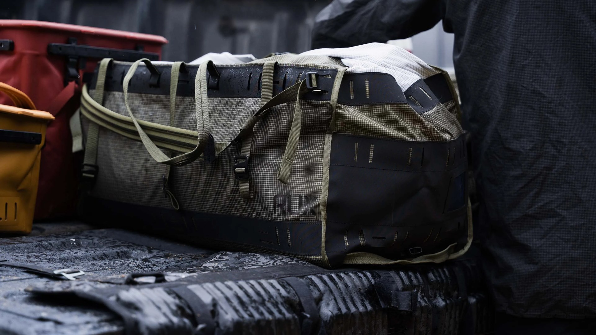 Large olive green and black RUX duffel bag with straps on a wet truck bed.