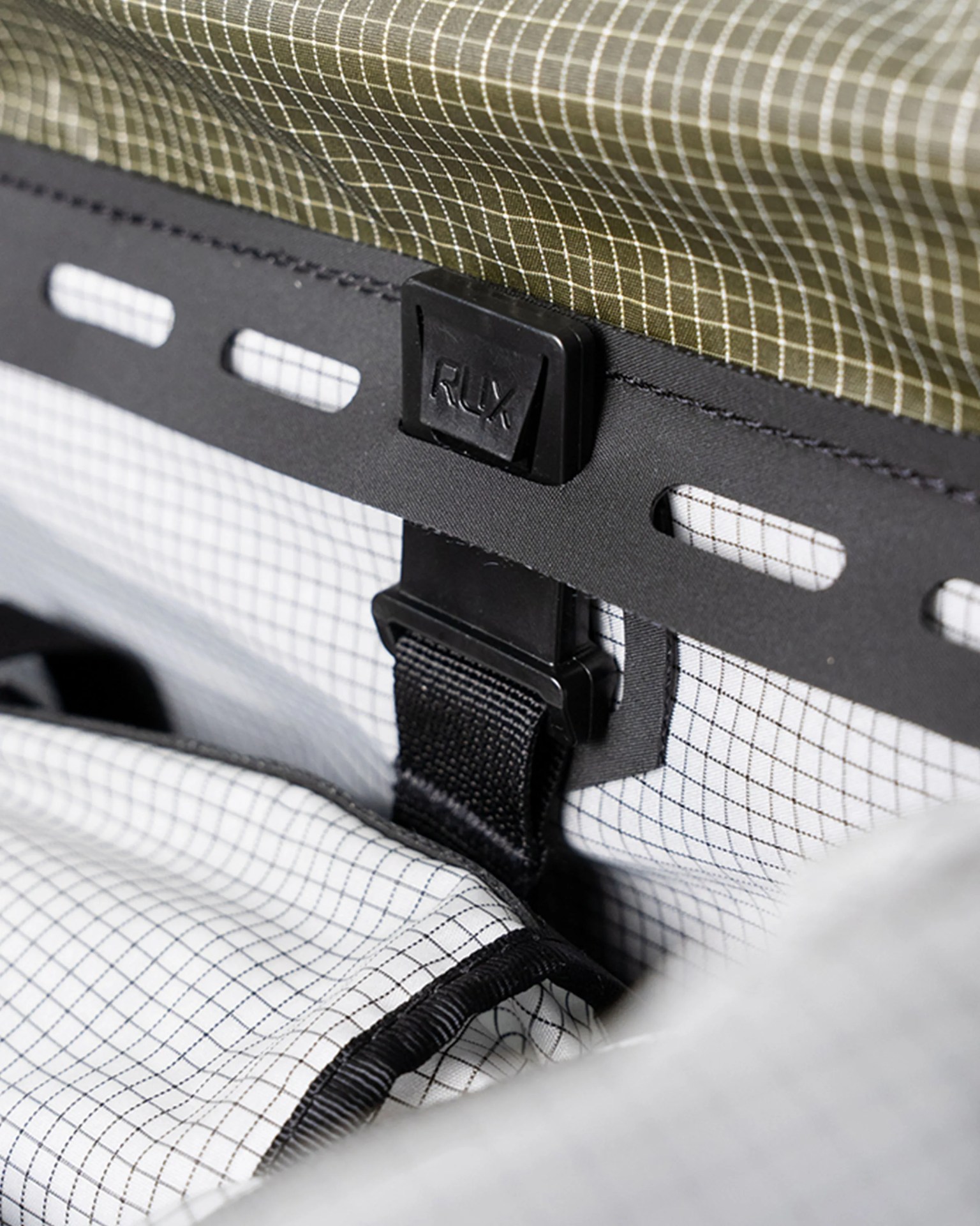 Close-up of a black plastic buckle with "RLX" embossed, attached to a black strap on white and olive grid-patterned fabric.