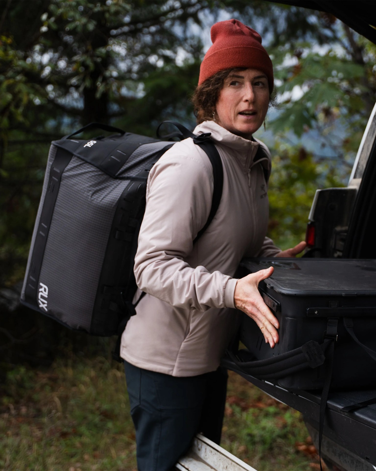 Person wearing a red beanie and beige jacket carrying a large gray and black RUX backpack while loading a black case into a vehicle.