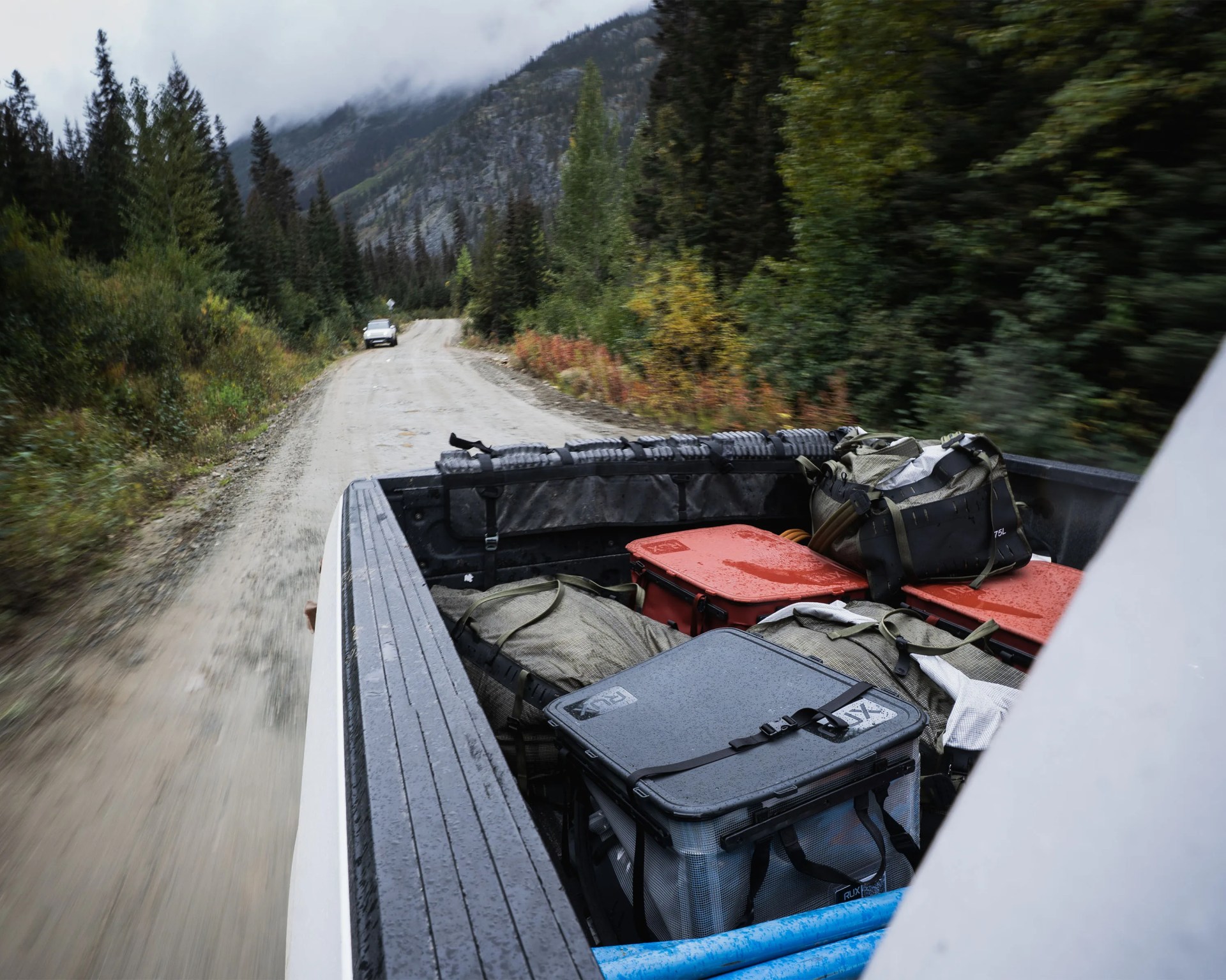 Pickup truck bed loaded with waterproof gear bags and cases on a forested dirt road.
