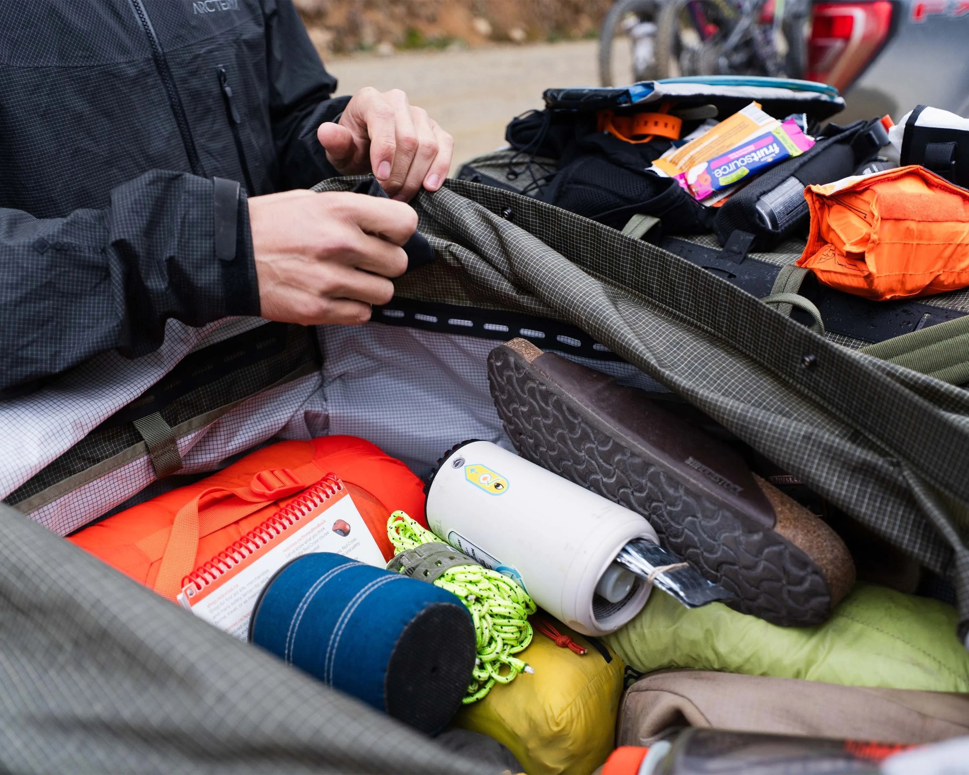 Hands opening a green checkered duffel bag filled with outdoor gear including a water bottle, rope, and orange bag.