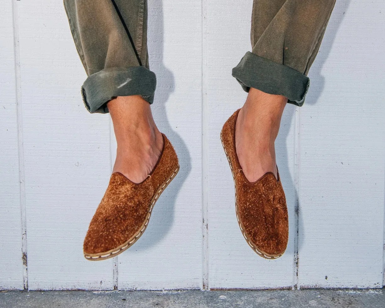 Brown textured slip-on shoes worn with rolled-up olive green pants against a white wooden wall.