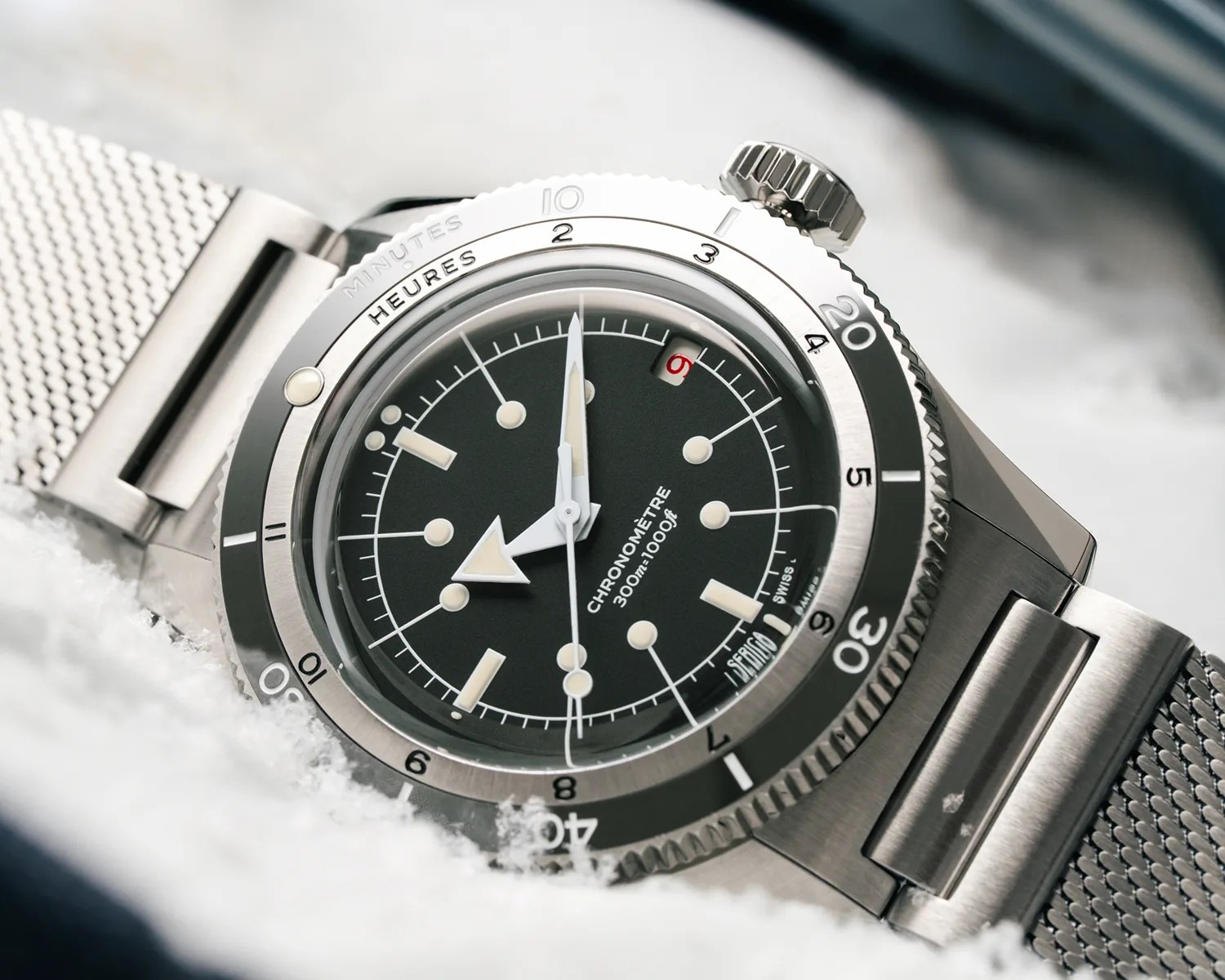 Stainless steel chronometer watch with black dial, white markers, and mesh bracelet resting on snow.