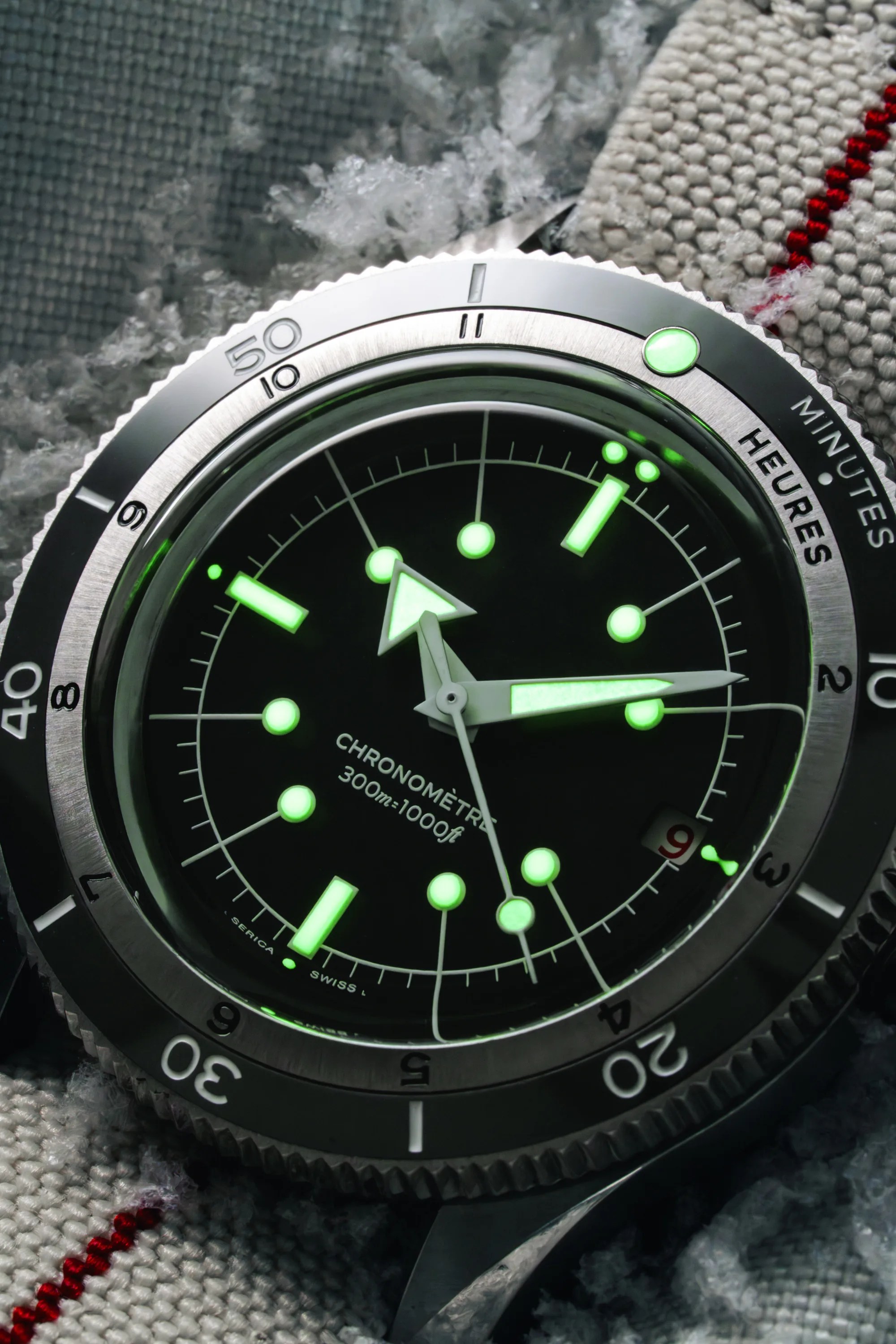 Close-up of a black dial chronometer watch with glowing green hour markers and hands on a fabric strap.