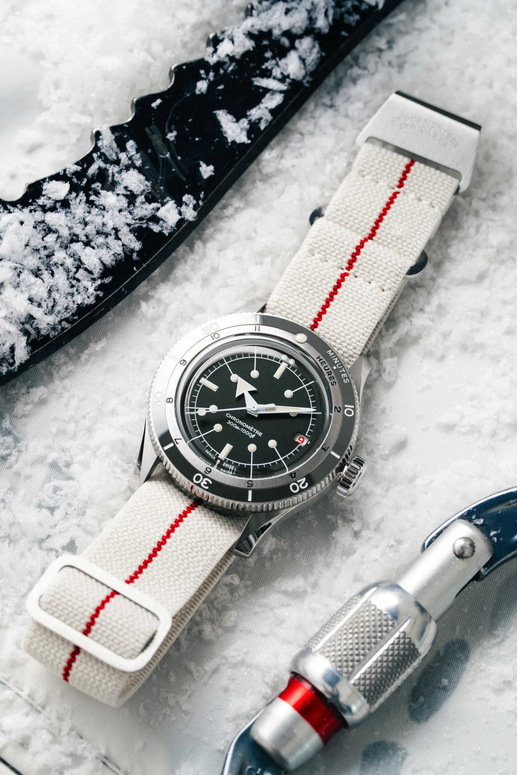 Silver dive watch with black dial and white canvas strap with red stripe on snow next to climbing gear.