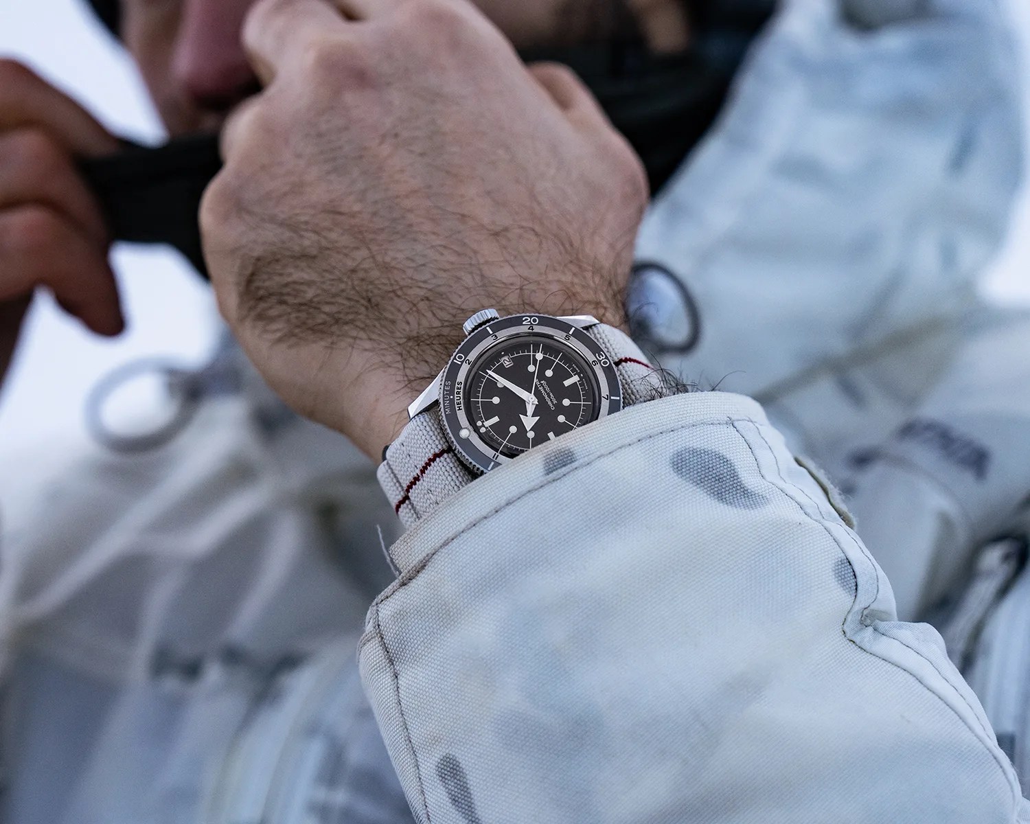 Black dial wristwatch with white markers and a gray fabric strap worn under a light gray jacket sleeve.