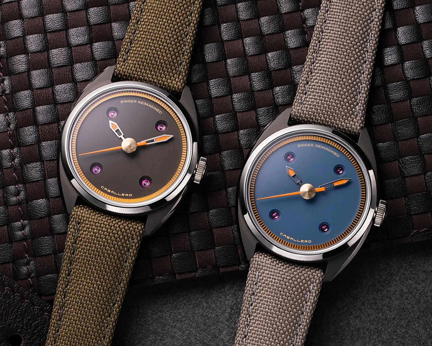 Two wristwatches with canvas straps, one with a brown dial and olive strap, the other with a blue dial and gray strap, on a woven leather surface.